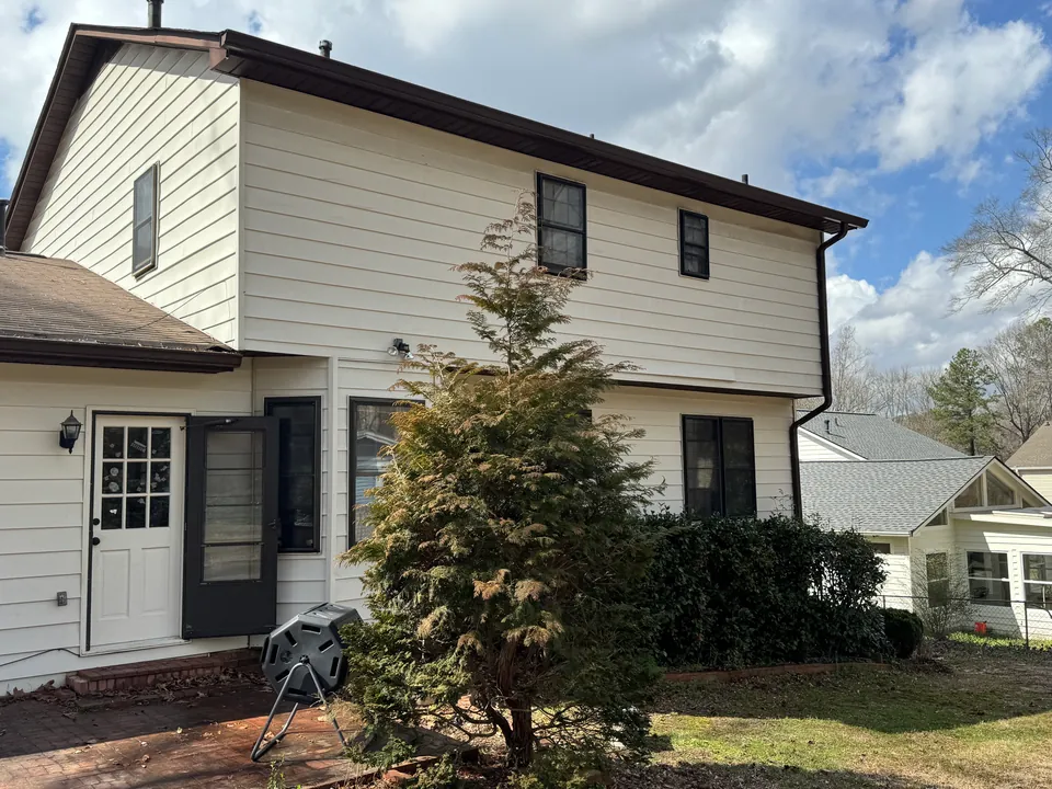 Cream two-story house exterior with dark trim, a lawn, shrubs and a patio beneath a partly cloudy sky.